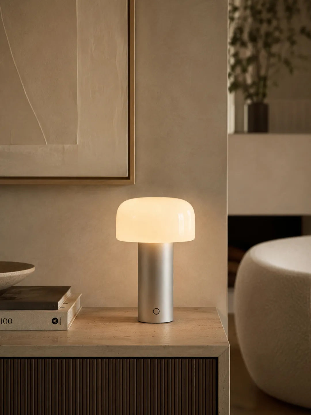 Hooled Vesper Metal Tischleuchte leuchtet auf einem Sideboard neben Büchern und einem Sessel
