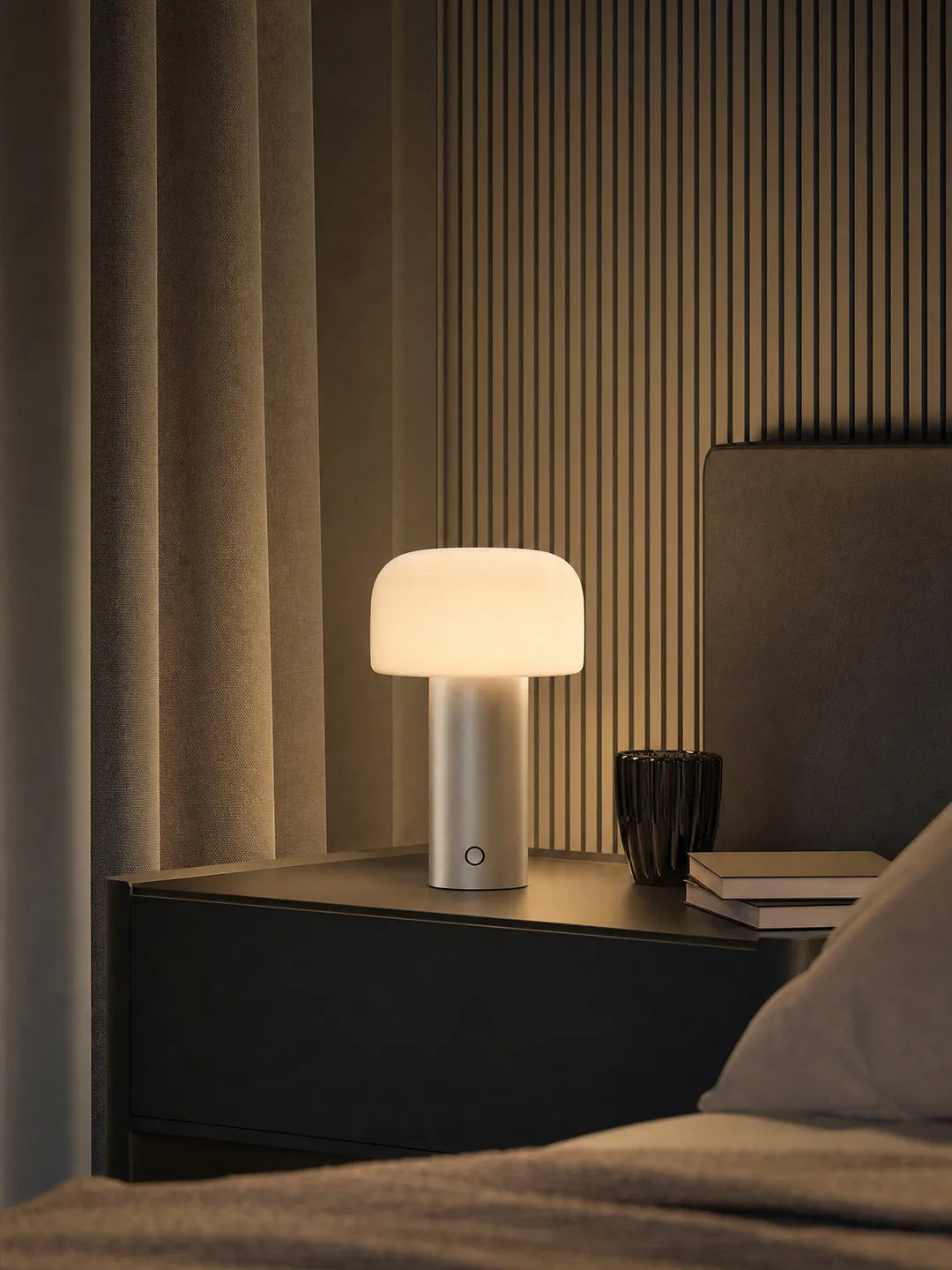 Hooled Vesper Metal Tischleuchte mit warmem Licht auf einem Nachttisch neben Bett und Büchern